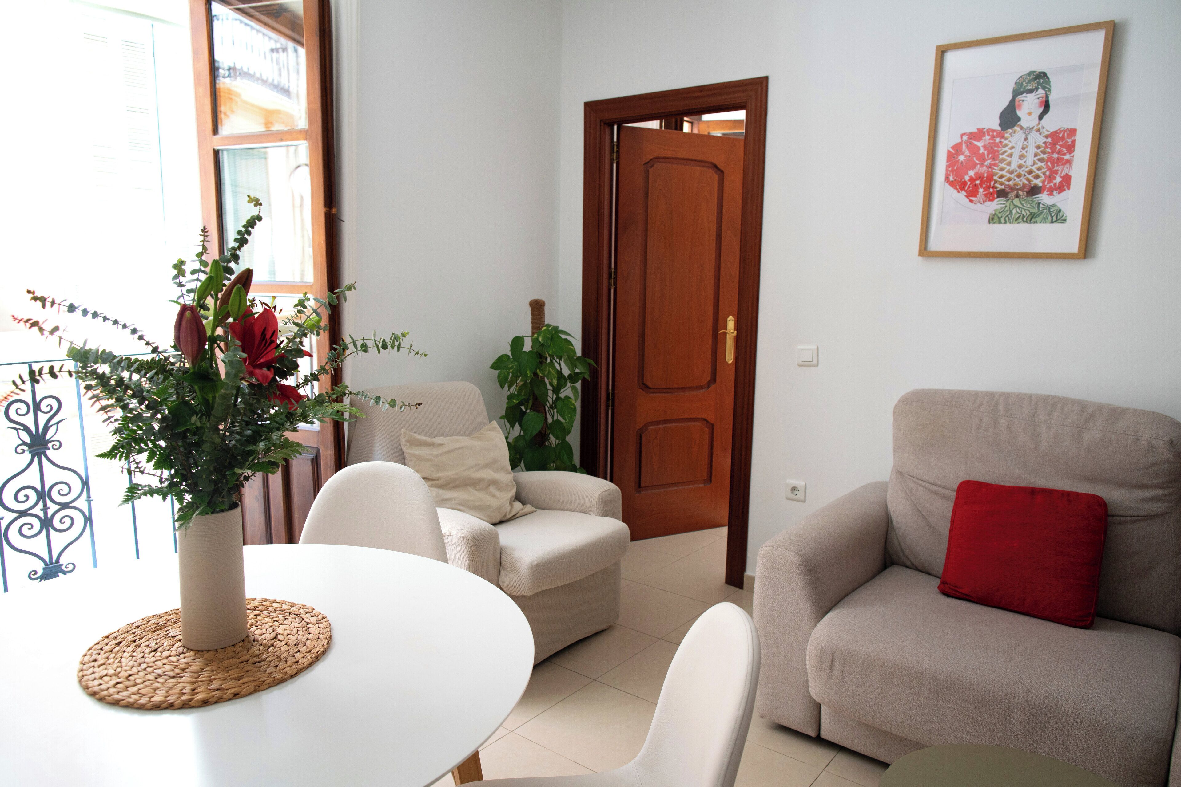 Un salón con una mesa blanca, sillones color beige, un cojín rojo y una puerta de madera.