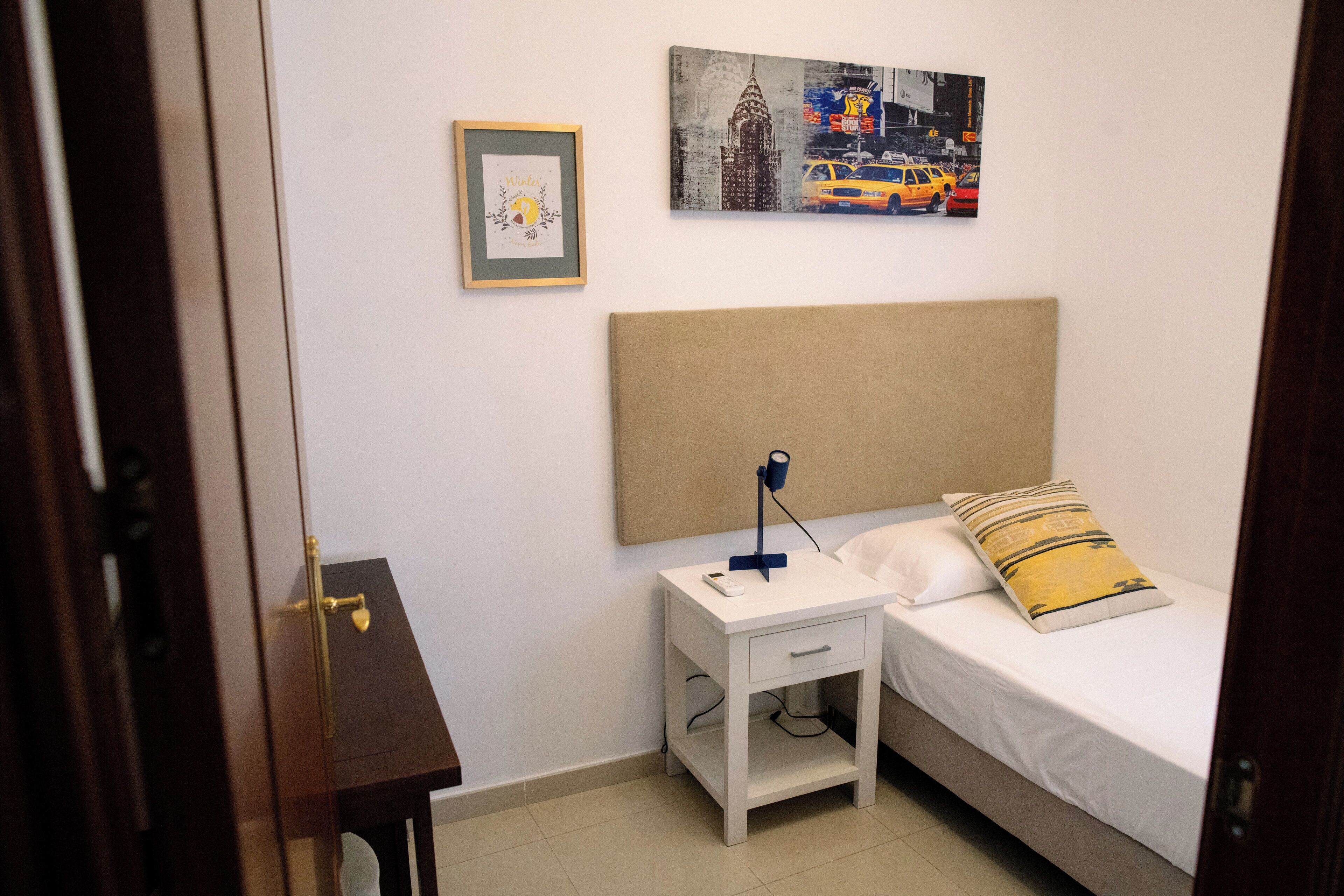 Un dormitorio con cama, mesita de noche y velador. Hay un cuadro en la pared y un cuadro sobre la cama.