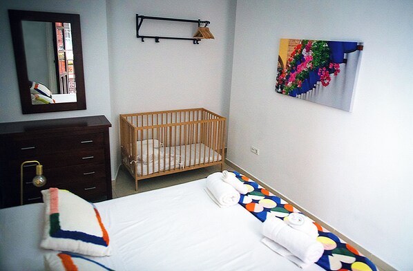 A bedroom with a crib, a bed with a colorful quilt, a dresser, a mirror, and a wall-mounted shelf. - El Marqués San Juan 1 (Málaga)