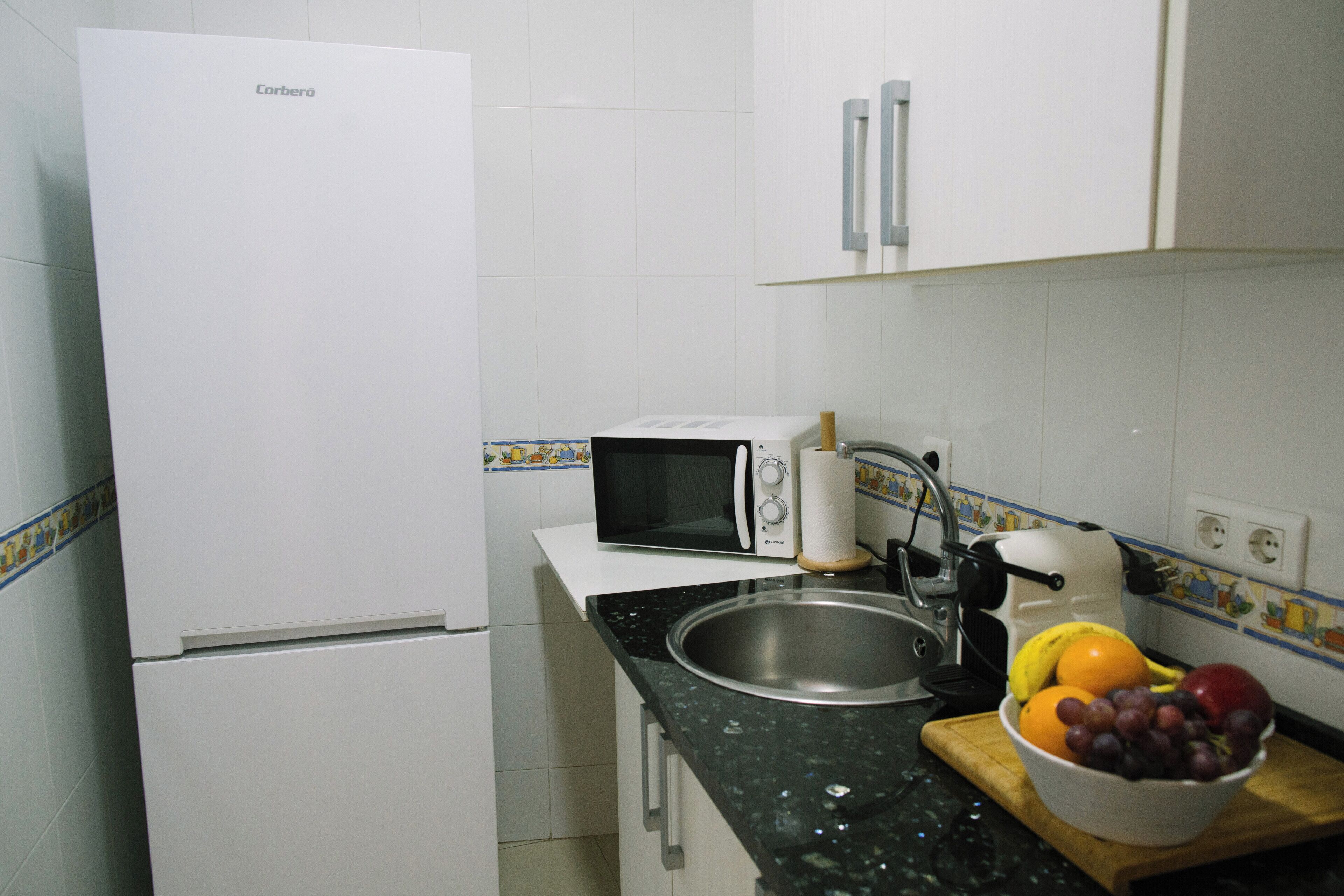 A compact kitchen with a white refrigerator, a microwave, a sink, and a bowl of fruit on the counter.