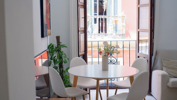 Un coin repas avec une table ronde et quatre chaises, une plante en pot, et une vue sur un balcon doté d’une balustrade et d’un immeuble en arrière-plan.
