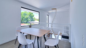 Un espace repas moderne avec une table en bois, des chaises blanches, une grande fenêtre donnant sur un paysage verdoyant et un luminaire suspendu.