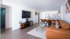 Un salon moderne avec un canapé en cuir, une table basse en verre et un téléviseur à écran plat posé sur un meuble en bois. On aperçoit à l’arrière-plan un coin repas avec une table et des chaises.