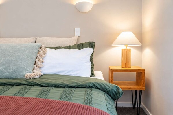 A neatly made bed with a green and white duvet, a wooden bedside table with a lamp, and a wall-mounted light fixture.