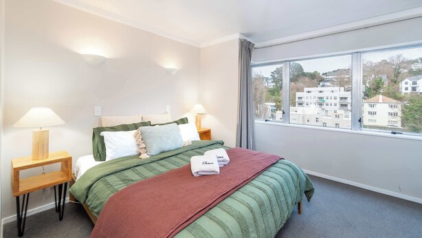 A hotel room with a bed, two bedside tables with lamps, a window with a city view, and a green and red blanket on the bed.