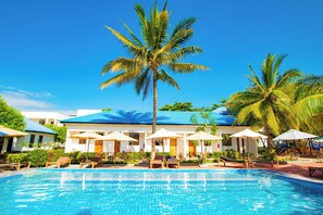 A swimming pool with lounge chairs and umbrellas, surrounded by palm trees and a building with a blue roof. - Kigwedeni Apartments (Nungwi)
