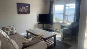 Een woonkamer met een bank, een salontafel, een tv op een meubelstuk en een raam met uitzicht op de zee.