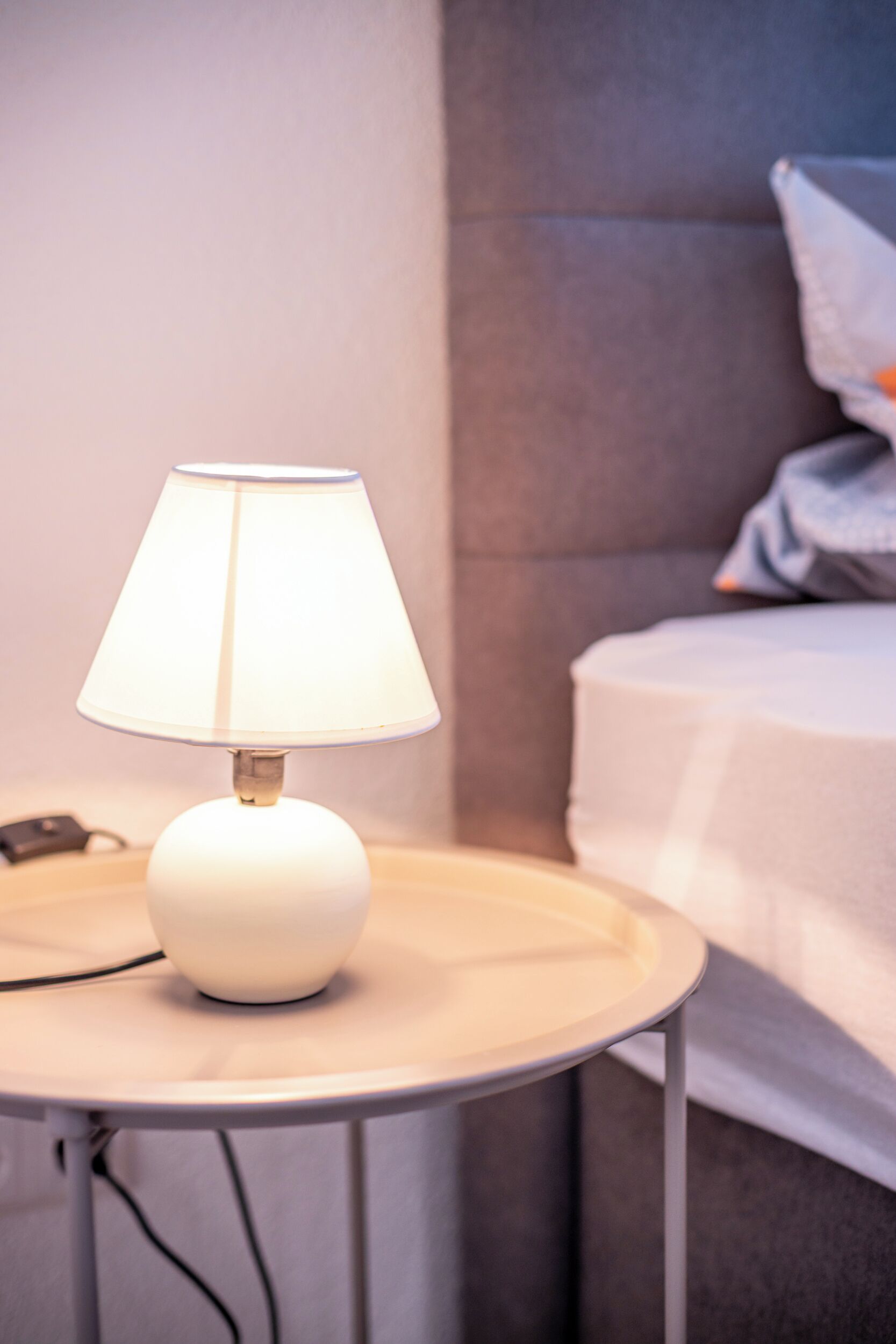 A bedside table with a white lamp, a bed with grey bedding, and a grey headboard.