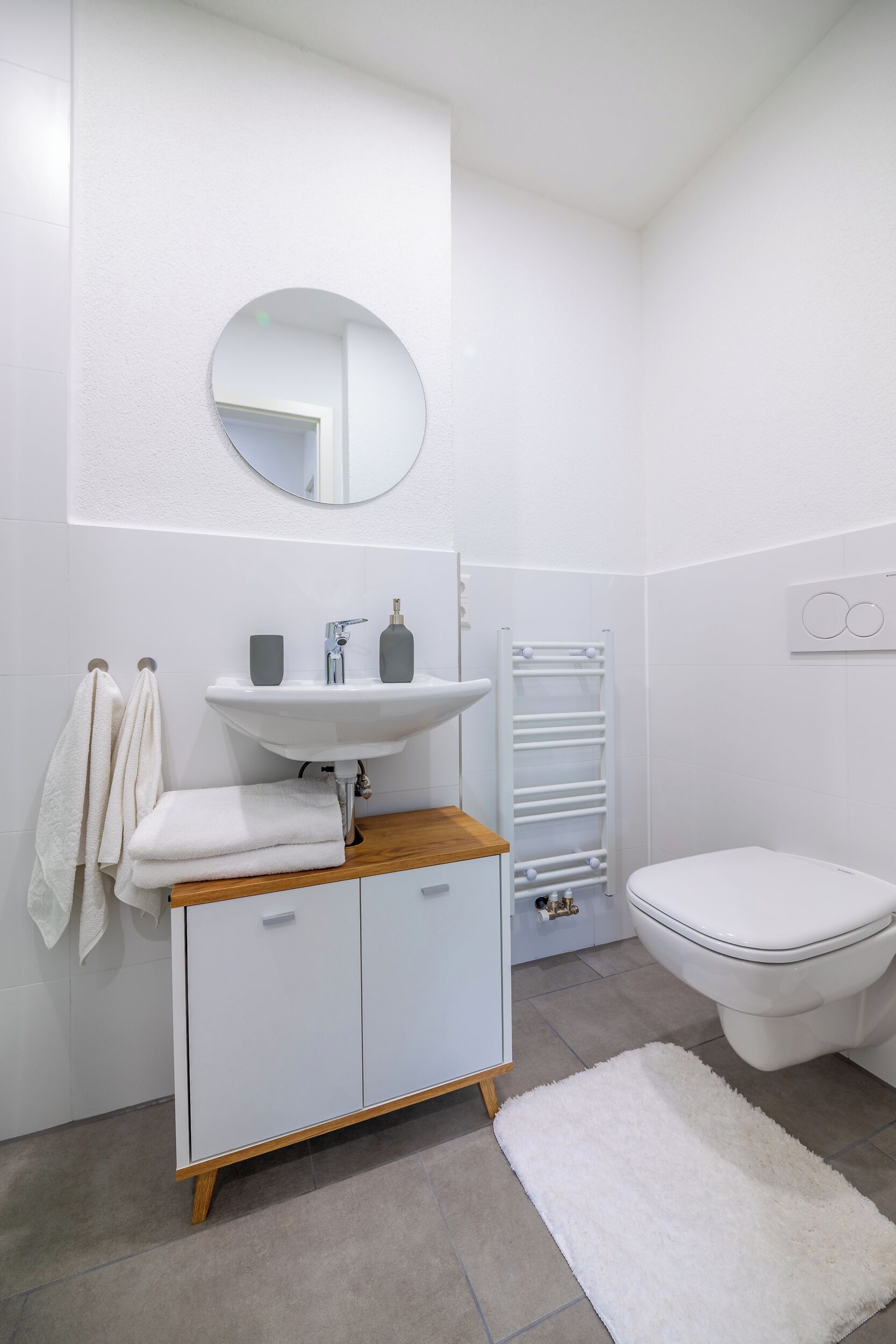 Un baño con lavamanos, espejo e inodoro blancos. Hay un perchero con toallas, una alfombra blanca y un mueble blanco con cajones.