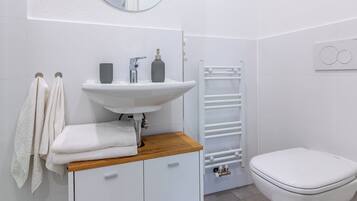 Un baño con lavamanos, espejo e inodoro blancos. Hay un perchero con toallas, una alfombra blanca y un mueble blanco con cajones.