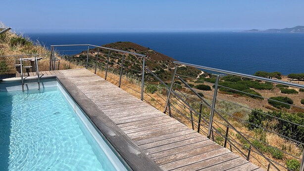 A wooden deck leading to a pool with clear blue water, overlooking a coastal landscape with trees and hills.