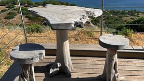 A wooden deck with two stone tables and a bench, overlooking a coastal landscape with mountains in the distance.