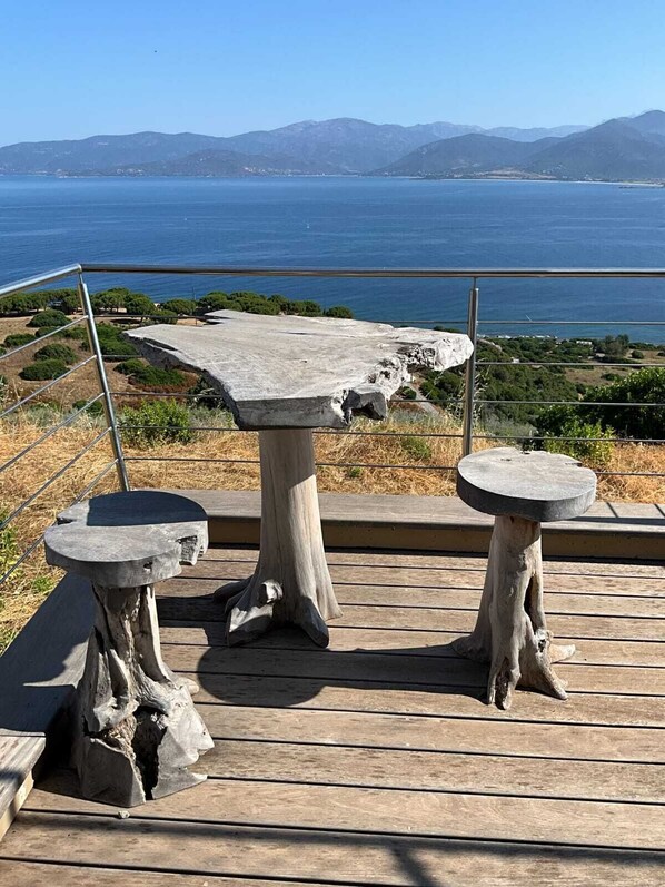 A wooden deck with two stone tables and a bench, overlooking a coastal landscape with mountains in the distance. - New apartment in a villa facing a panoramic sea view with sunsets over the sea (Appietto)