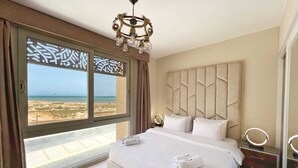 Ein Schlafzimmer mit einem großen Bett, einer Kristallleuchter, einem Schminktisch mit Spiegel und Blick auf den Strand durch ein Fenster.