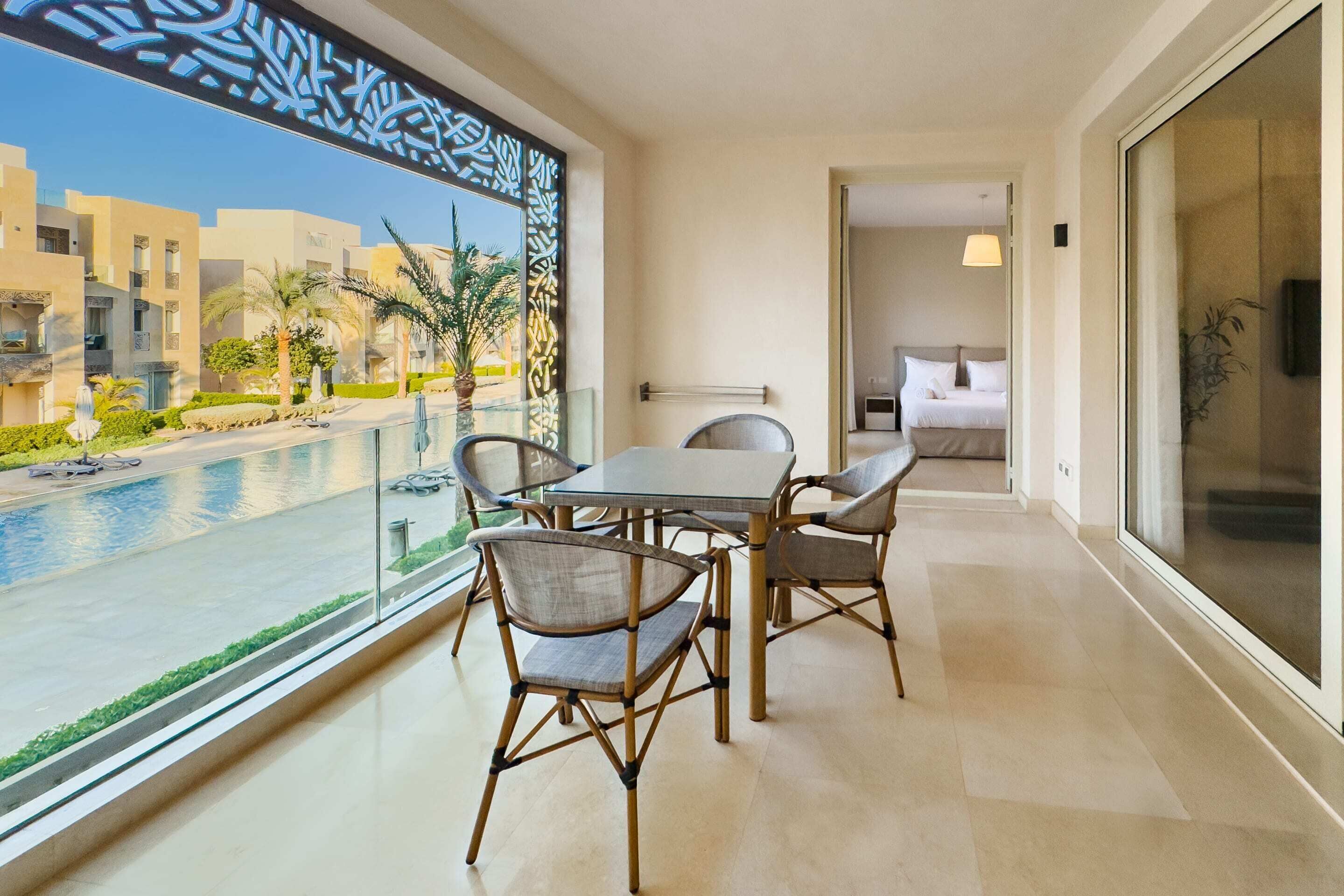 Un balcón con vistas a la piscina, una zona de comedor con sillas y un dormitorio con cama y mesita de noche.