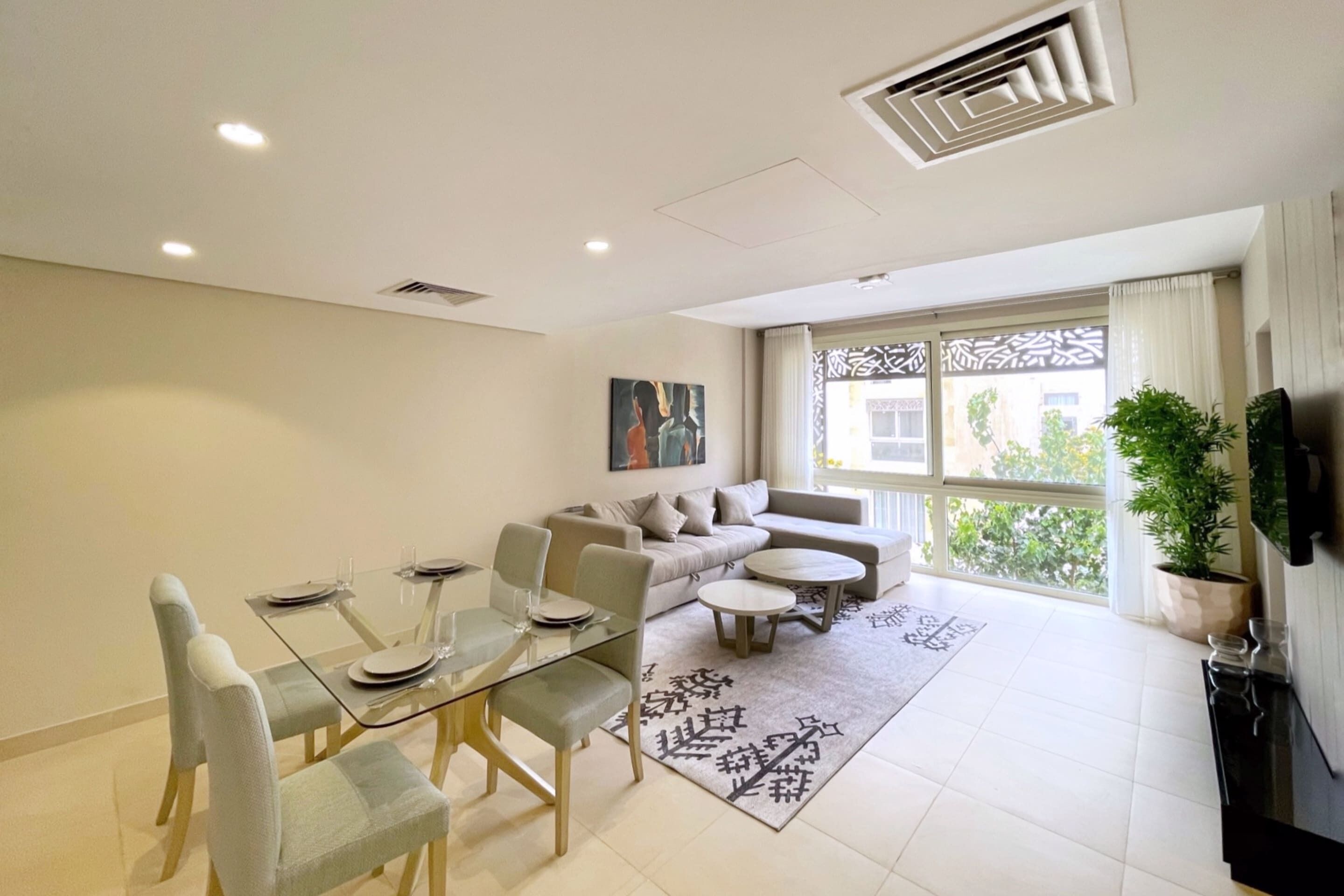 A modern living room with a glass dining table, sofas, and a large window with a view.