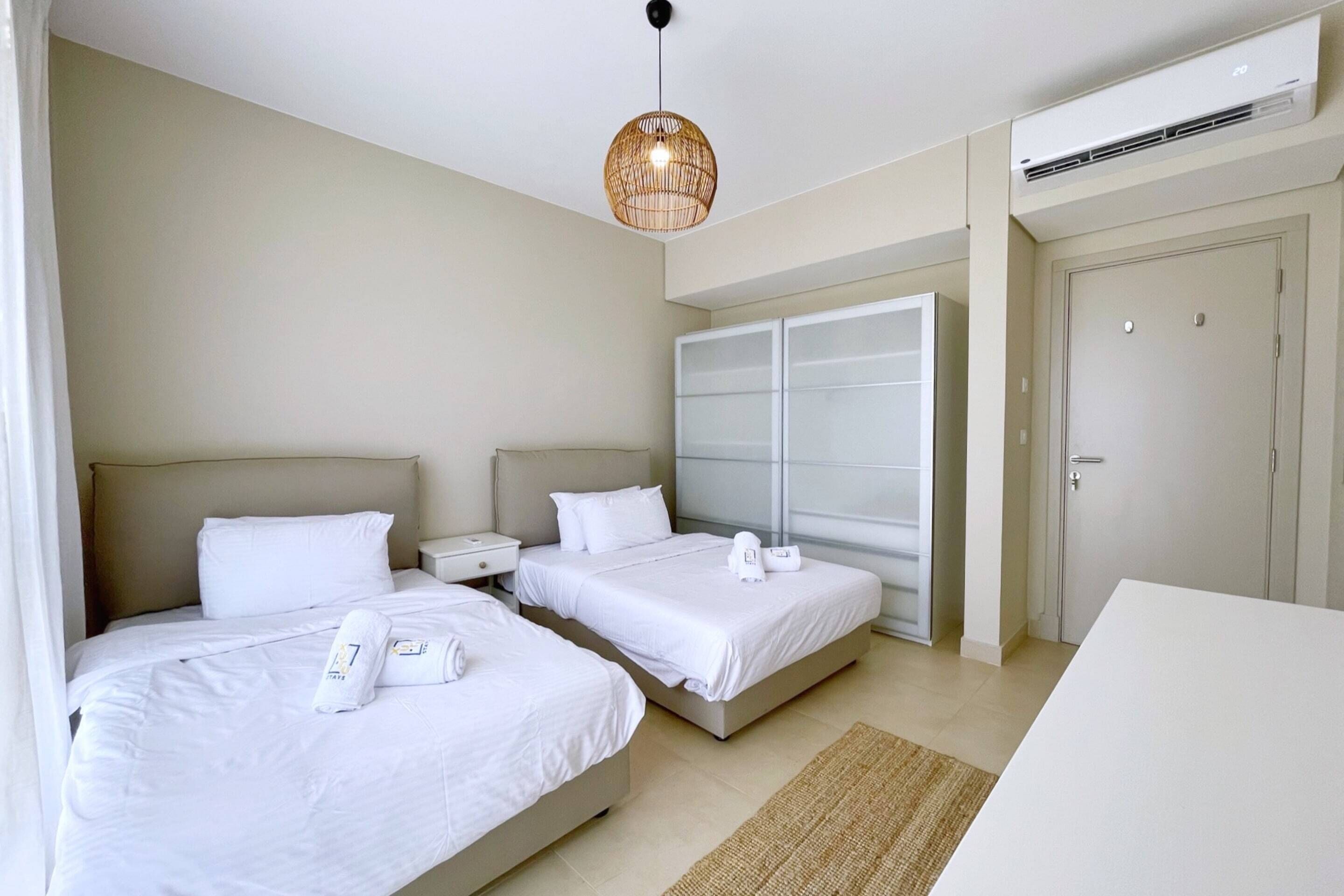 A hotel room with two beds, a bedside table, a wardrobe, and a hanging light fixture.