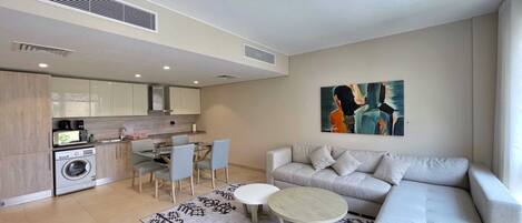 A modern living room with a grey sofa, a coffee table, and a dining area with chairs and a table. There is a kitchen area with a washing machine and a sink.