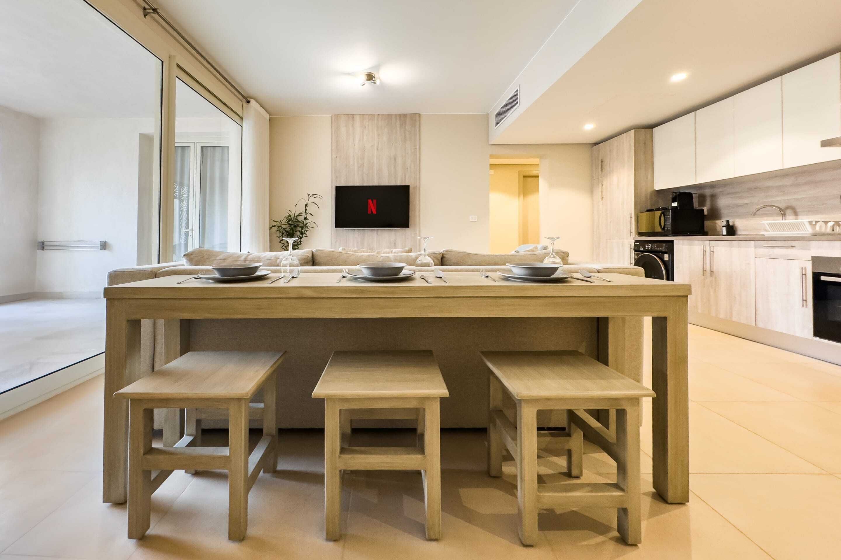A modern kitchen with a central island, wooden stools, and a flat-screen TV mounted on the wall.