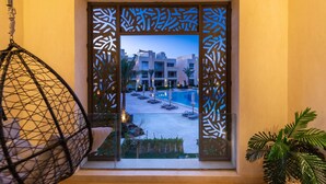 Vista a través de una puerta decorativa con patrones geométricos, que da a una zona de piscina con sillones y sombrillas, y un edificio con varios balcones.