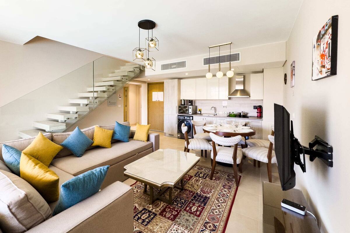 A modern living room with a sofa, coffee table, and patterned rug. There is a staircase leading to an upper level, a kitchen area with a dining table, and a mounted TV.