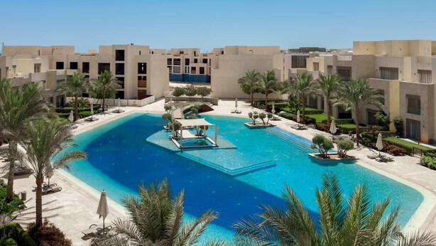 Un complejo turístico con piscina central, palmeras y edificios modernos.