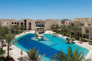 A resort with a central swimming pool, palm trees, and modern buildings. - Xuru Stays I Modish & Magnific 2bdr @ Mangroovy Pool & Beach Access (El Gouna)