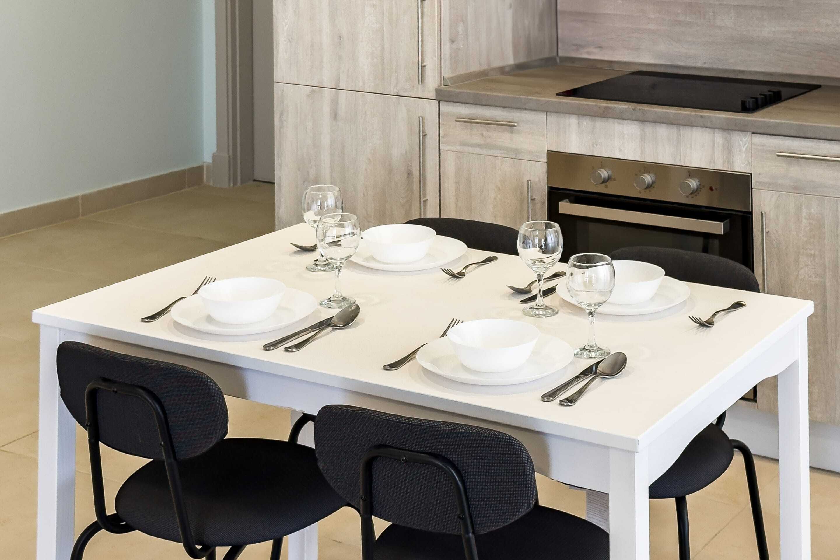 A dining table set for four with white plates, bowls, and cutlery, and black chairs.