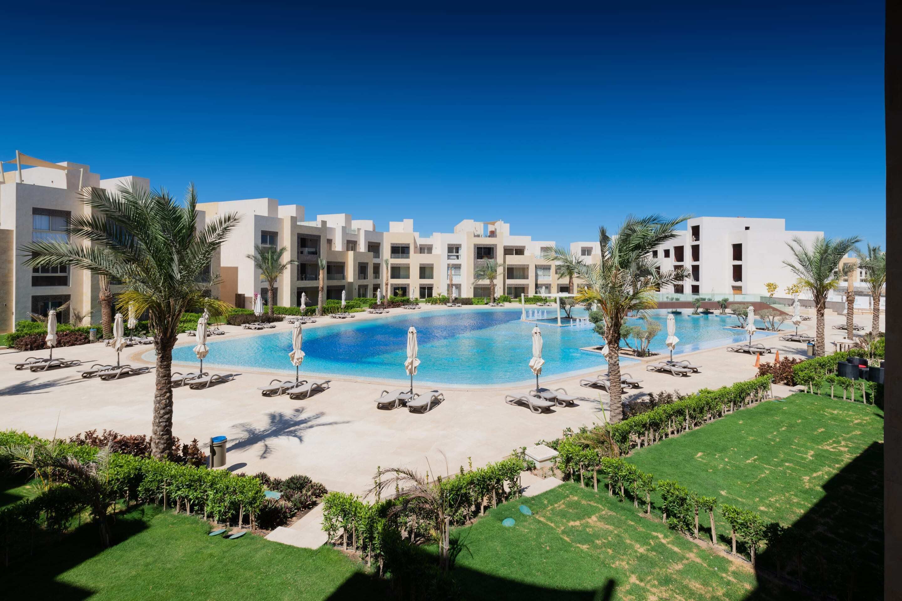 A hotel with a large swimming pool surrounded by sun loungers and palm trees.