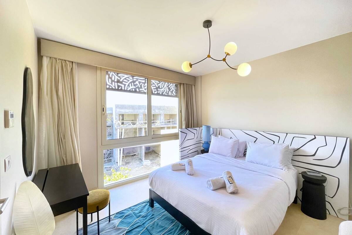 A hotel room with a large bed, a desk, a chair, a small stool, a trash bin, and a window with a view of buildings and greenery.
