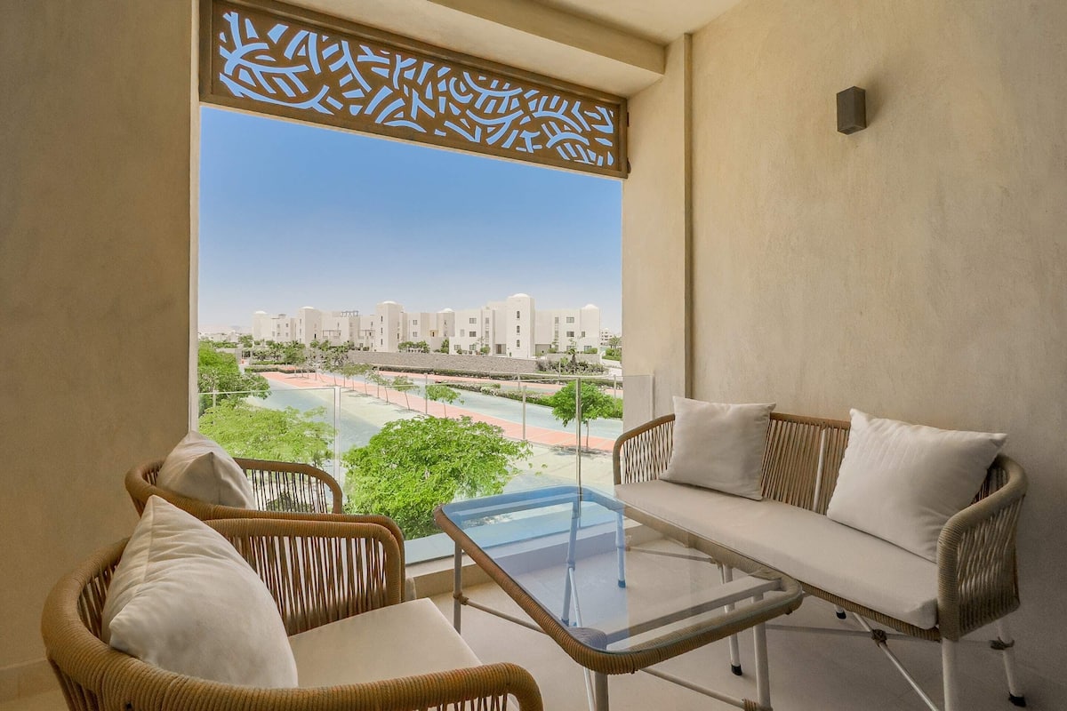 A balcony with wicker furniture, a glass table, and a view of a cityscape and greenery.