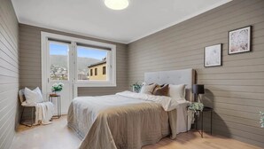 A bedroom with a large bed, a window with a view, a bedside table with a lamp, and a chair.