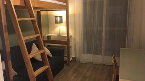 Une chambre avec un lit superposé, une échelle, un bureau, une chaise, une lampe et une fenêtre avec des rideaux légers.