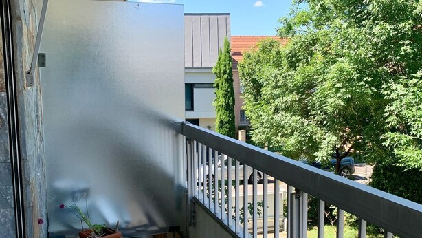 Un balcon orné d’un pot de fleurs, doté d’une rambarde en métal, et offrant une vue sur un immeuble et des arbres.