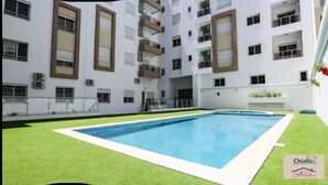 Une piscine entourée de gazon synthétique, avec un immeuble à plusieurs étages en arrière-plan.