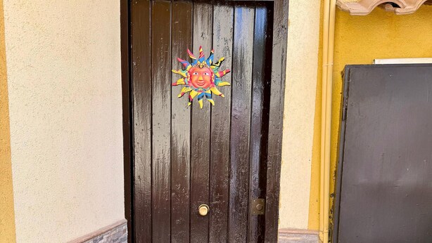 Una puerta de madera oscura con un emblema de sol decorativo, un felpudo y un acceso con suelo de piedra.