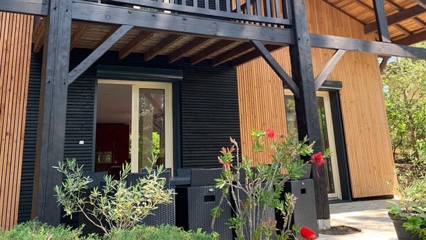 A two-story wooden house with a balcony, large windows, and a garden with various plants and flowers. - Rental of a villa with swimming pool, close to the lake and ocean beaches. (Lacanau)