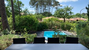 A deck with four chairs overlooking a swimming pool surrounded by lush greenery and tall trees. - Rental of a villa with swimming pool, close to the lake and ocean beaches. (Lacanau)