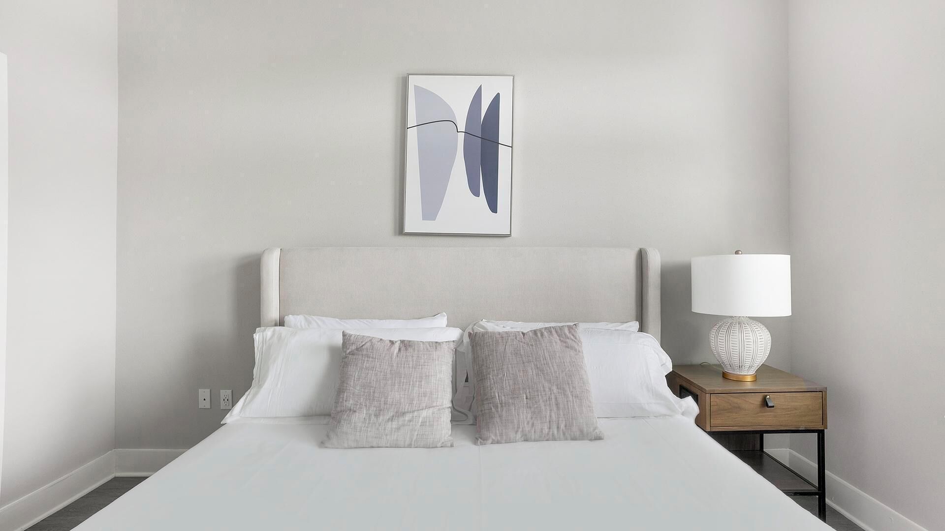 A neatly made bed with white and grey linens, a modern headboard, and a bedside table with a lamp.