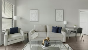 Una sala de estar moderna con un sofá, dos sillones, una mesa de centro de vidrio y un escritorio con una silla. Hay dos obras de arte abstractas enmarcadas en la pared.