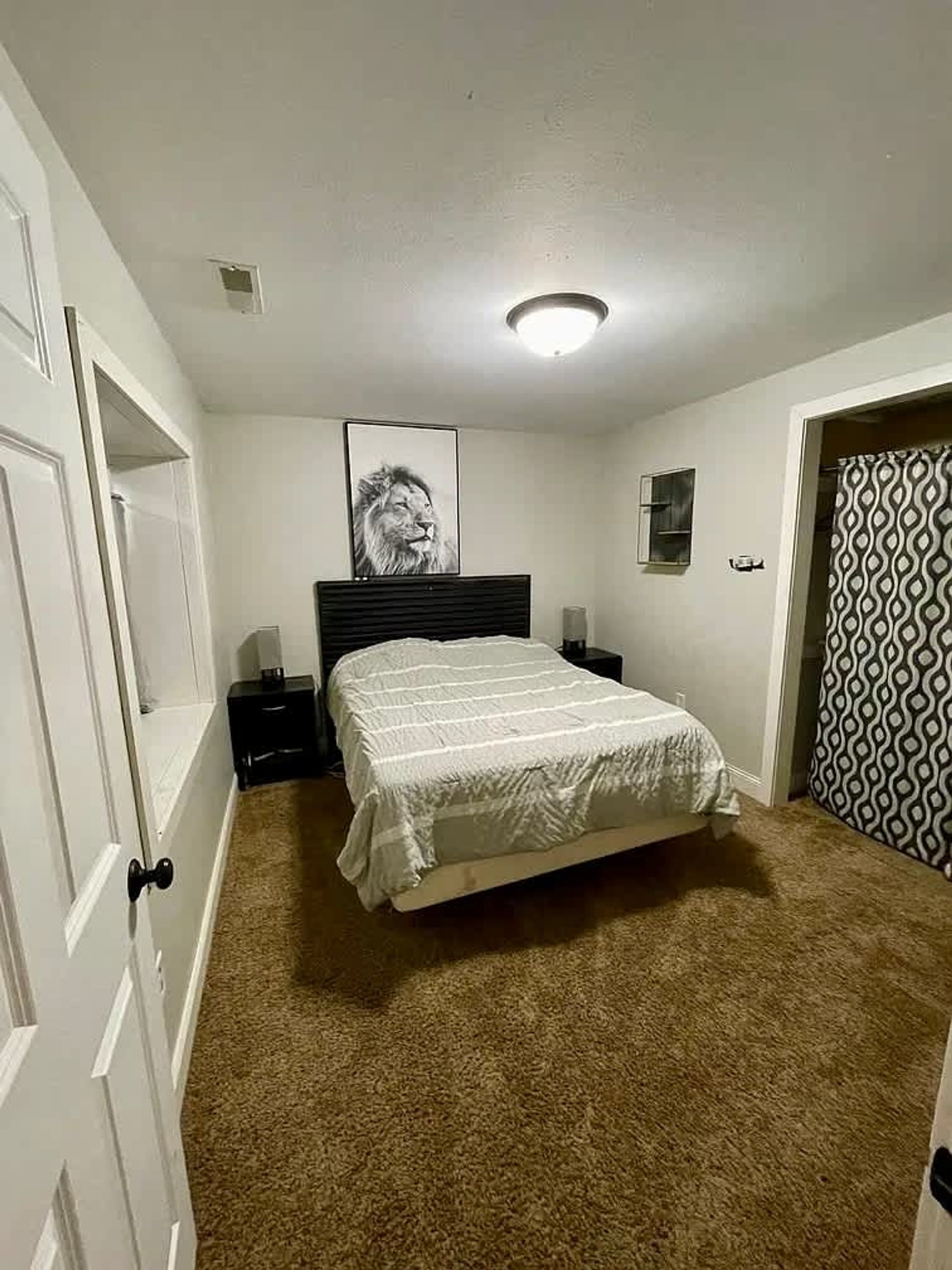 Een slaapkamer met een bed, een nachtkastje, een ingelijst beeld aan de muur en een deur met een patroonkordijntje.