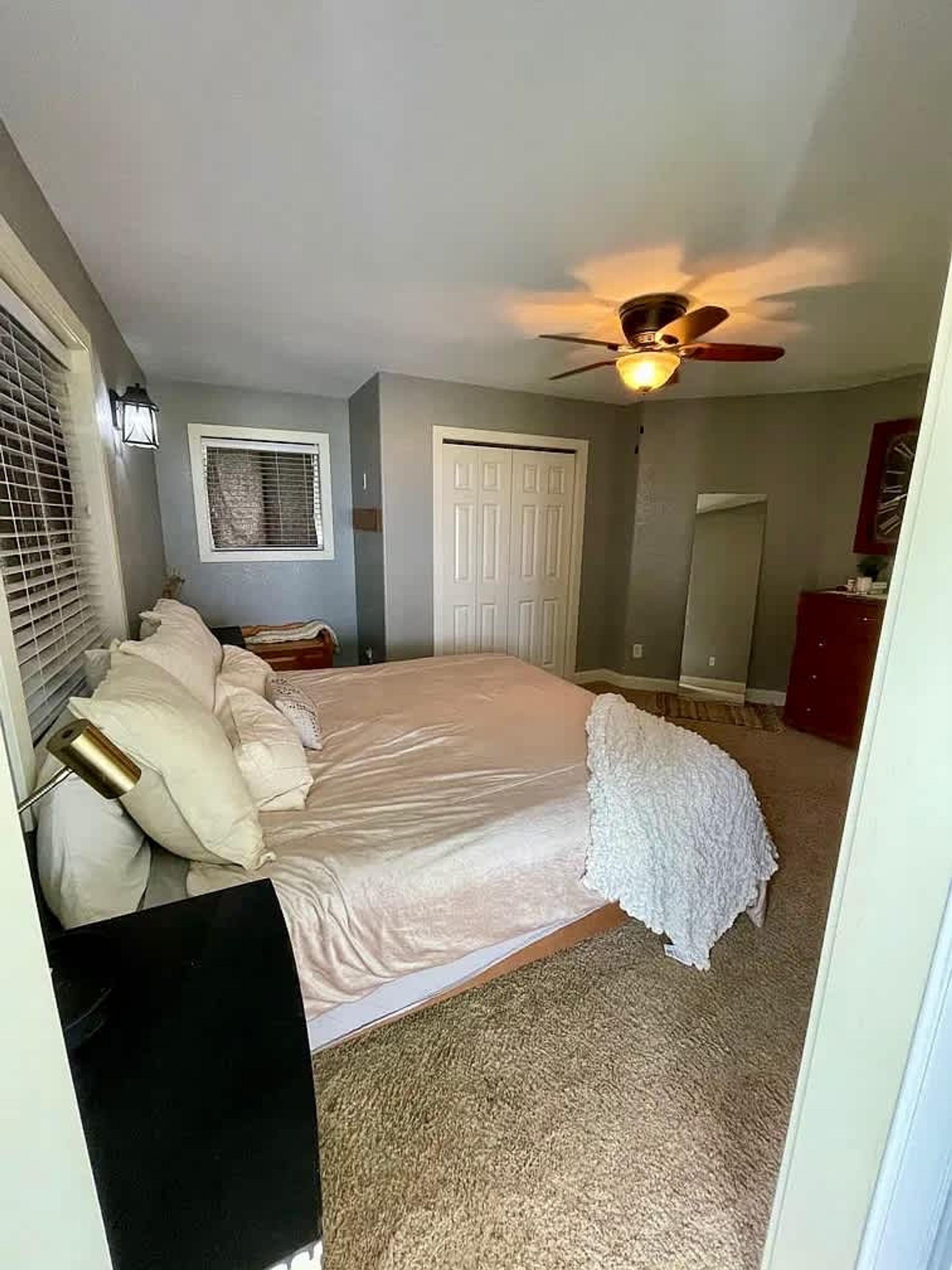 A bedroom with a bed, a ceiling fan, a window with blinds, a door, and a small dresser.