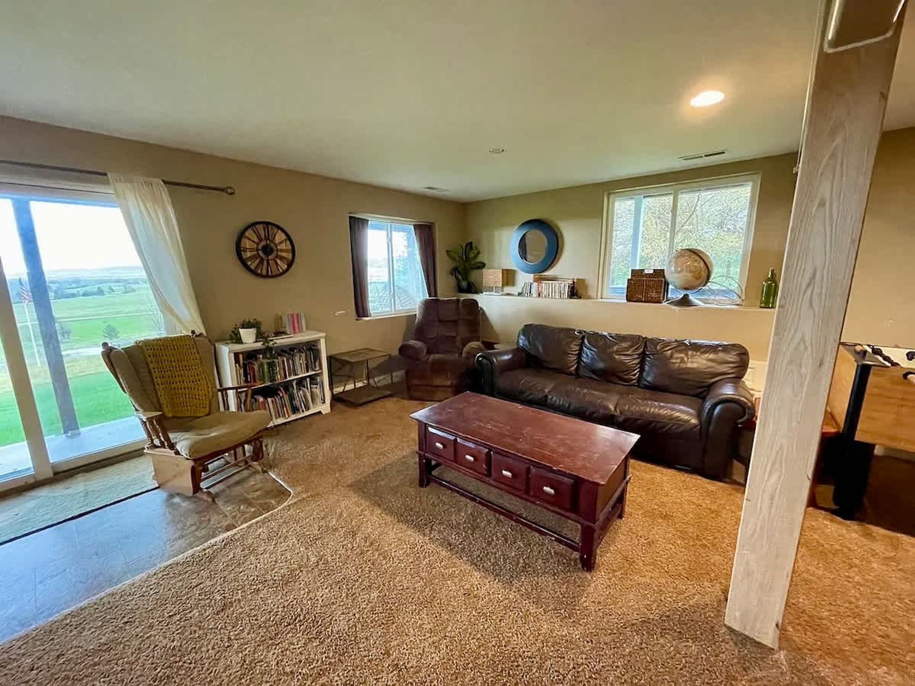 A cozy living room with a leather sofa, a wooden coffee table, and a bookshelf. There is a large window with curtains, a dartboard on the wall, and a view of the outdoors.