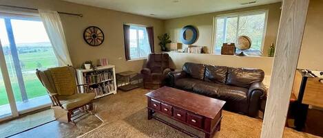 A cozy living room with a leather sofa, a wooden coffee table, and a bookshelf. There is a large window with curtains, a dartboard on the wall, and a view of the outdoors.