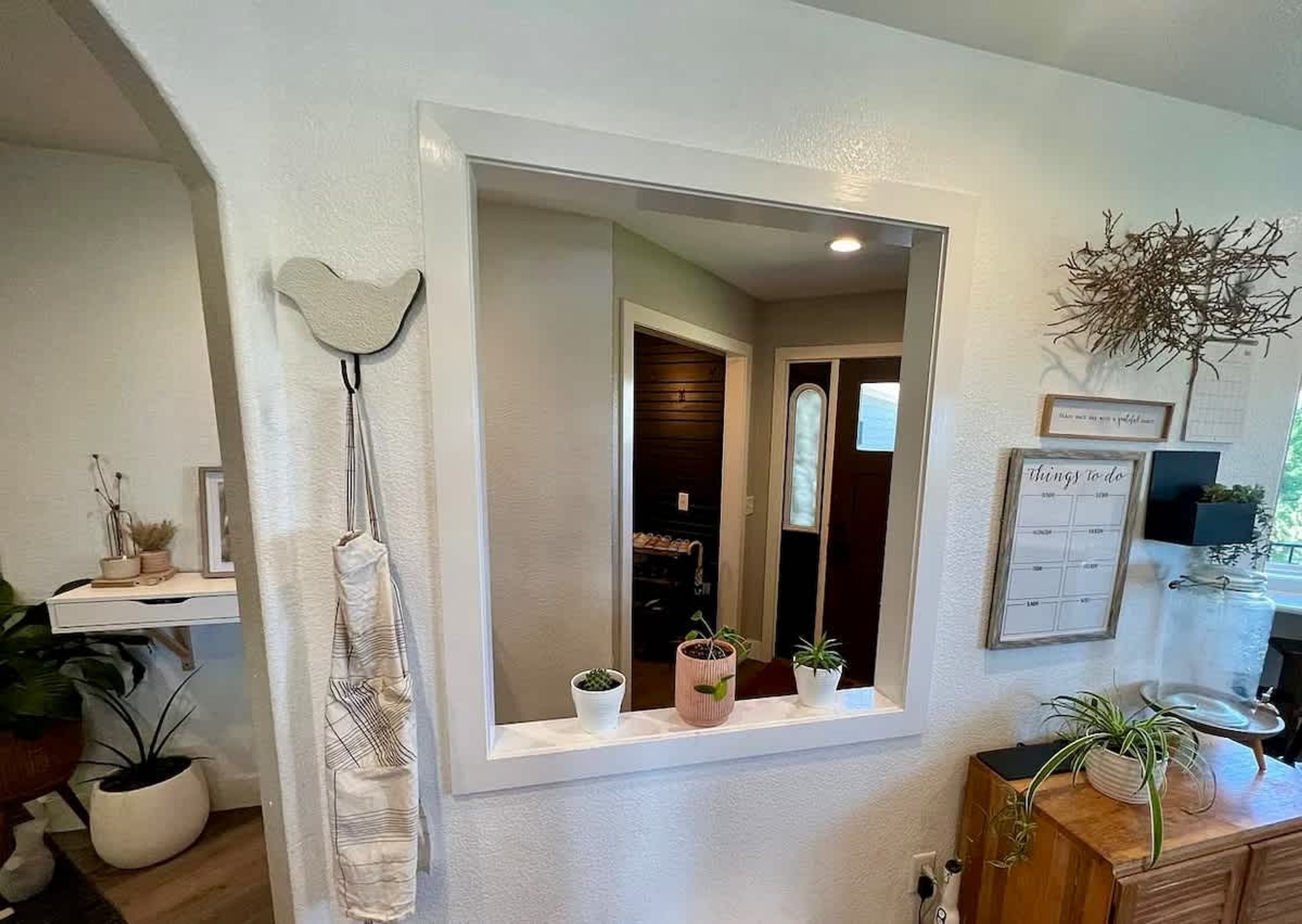 A room with a large mirror, a window with potted plants, a wooden cabinet, and a wall-mounted shelf with decorative items.