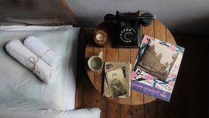 Ein ordentlich bezogenes Bett mit aufgerollten weißen Handtüchern, ein altes Wählscheibentelefon, eine Tasse und ein Fotoalbum auf einem Holztisch in einem Zimmer mit Holzboden.