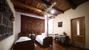 Ein traditionell eingerichteter Raum mit zwei Holzbetten, einer Steinwand, einem großen, gemusterten Wandteppich und einer Holztür mit Glasfeldern.