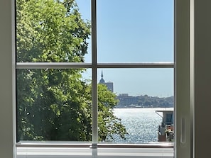 Une vue à travers une fenêtre, avec un ciel dégagé, une étendue d’eau et un bâtiment éloigné surmonté d’une flèche.