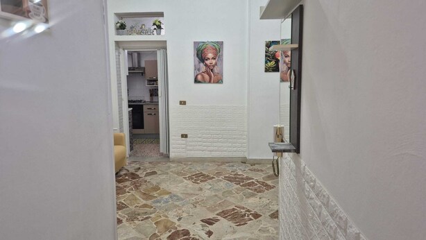 A narrow hallway with a tiled floor, a kitchen visible through an open door, and a small shelf with decorative items. - Little Brazil Home (Capaci)