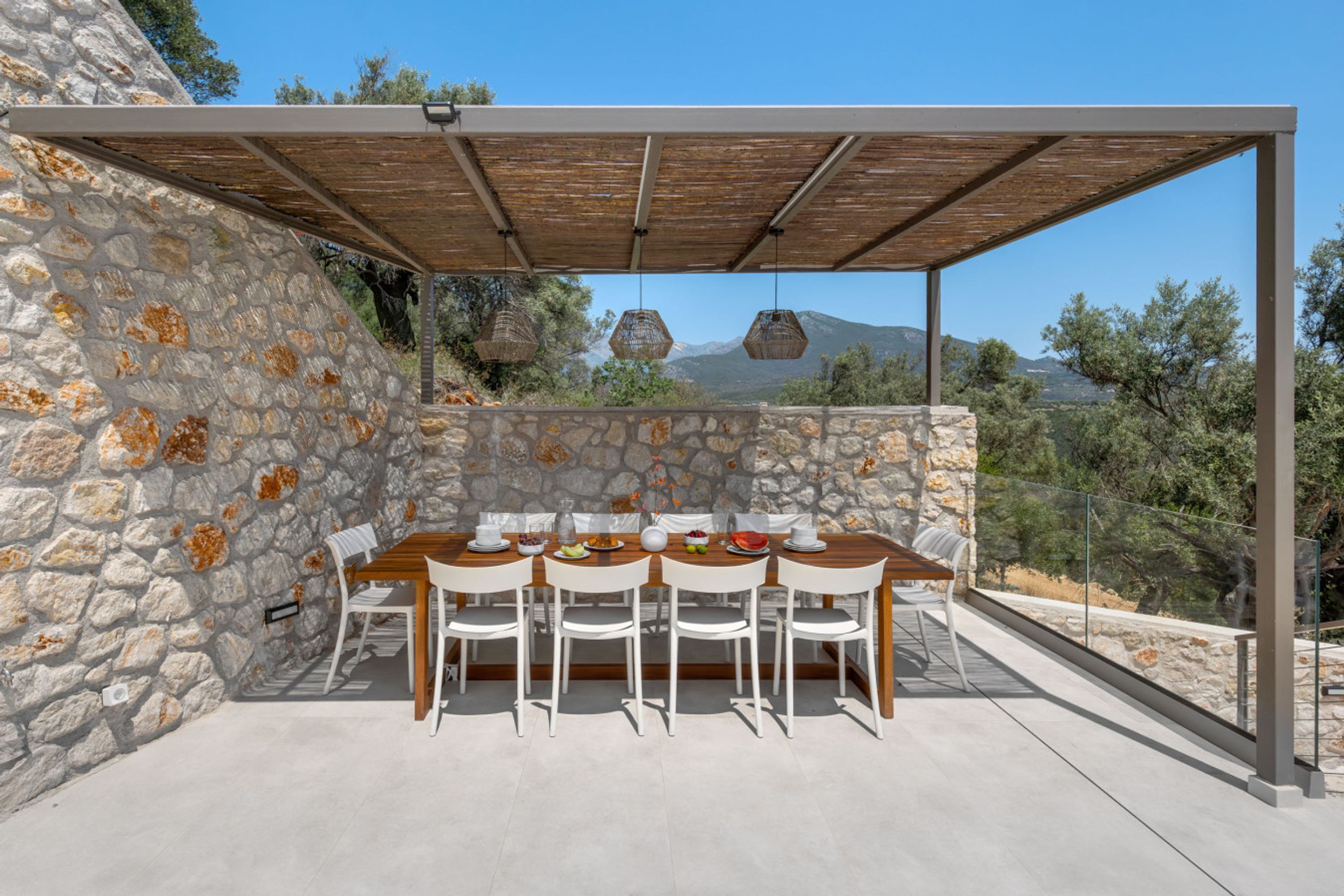 An outdoor dining area with a stone wall, wooden table, white chairs, and hanging light fixtures.